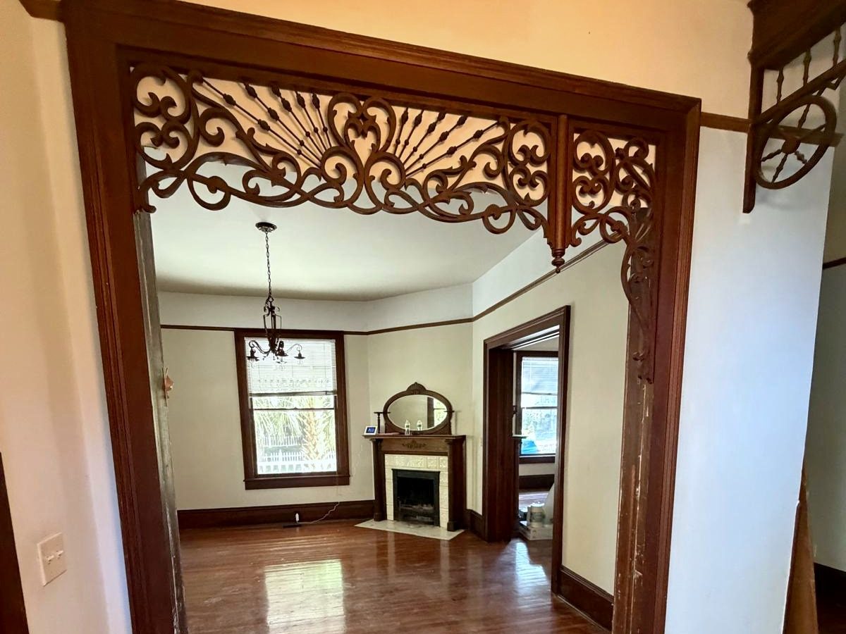 Looking through a carved fretwork archway into the living room: wide-plank wood floors, dark-stained trim, a chandelier and a wood mantel with a tiled fireplace and an oval mirror.