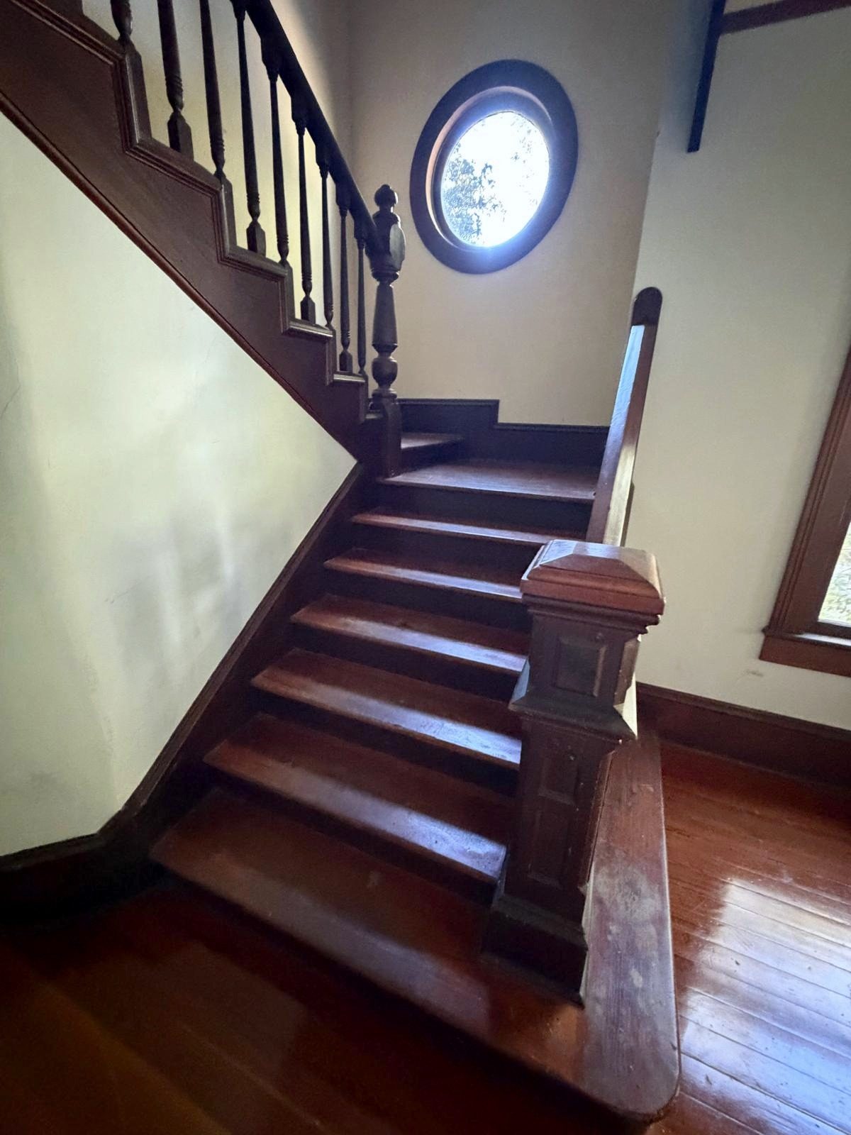 The staircase turning up past a round window, with turned balusters, a paneled newel post and wood treads.