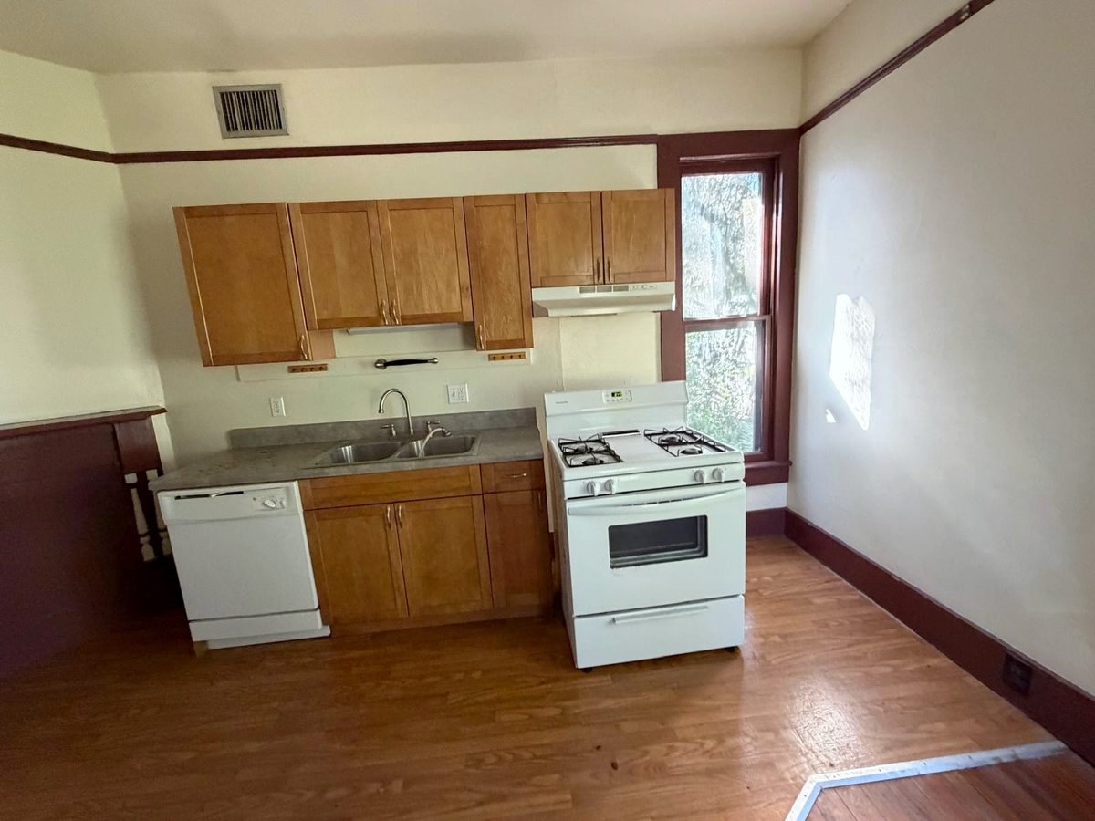 The kitchen before the work: honey-colored cabinets, laminate counters, a white gas range and dishwasher, and laminate flooring laid over the original wood.